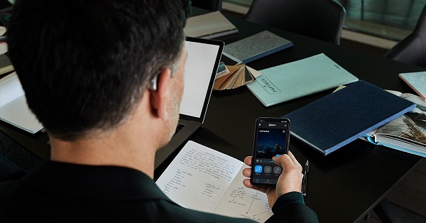  20.01-Widex-MOMENT-Office-Man-sitting-at-desk-seen-from-back-smartphone-in-hand 1
